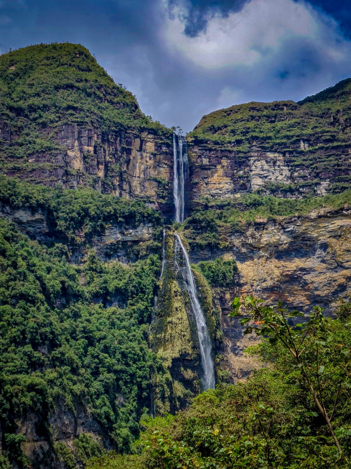 Cascade majestueuse tombant d'une falaise verdoyante sous un ciel nuageux.